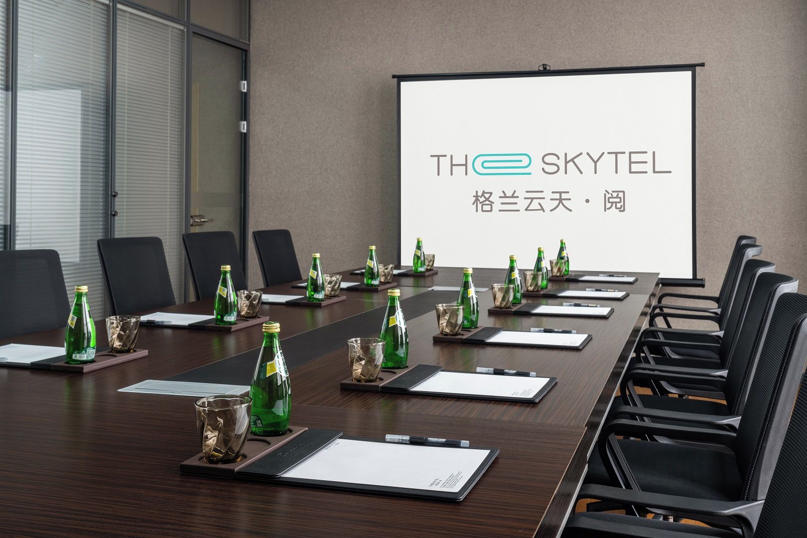 A conference room with a long table, chairs, a projector screen, and green bottles on coasters.