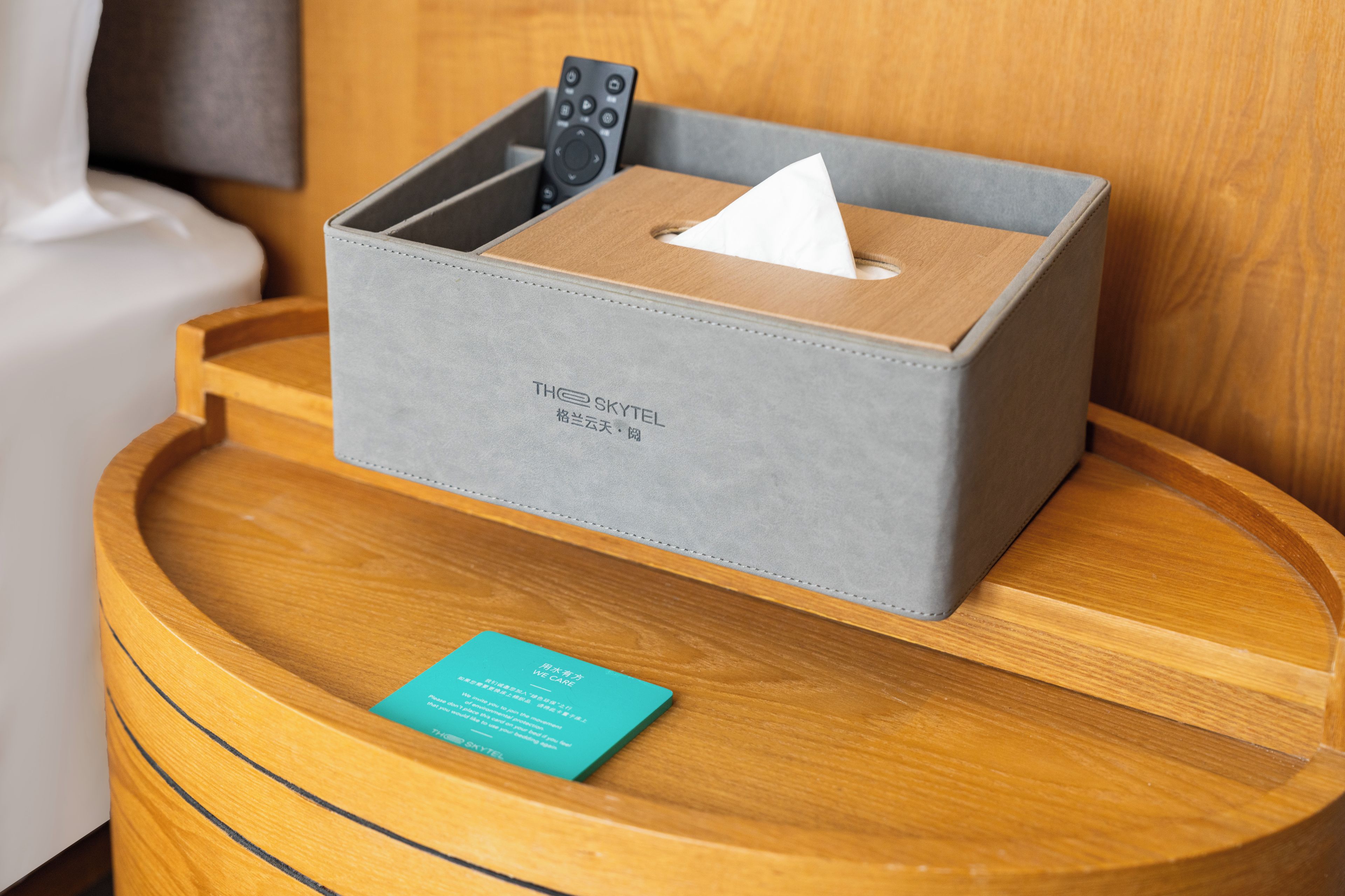 A grey box with a remote control and a tissue on a wooden bedside table.