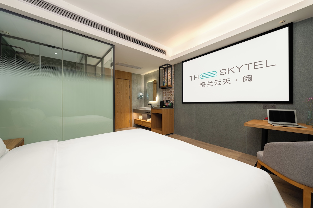 A modern hotel room with a large bed, a desk with a laptop, a shower area, and a large screen displaying the hotel name.