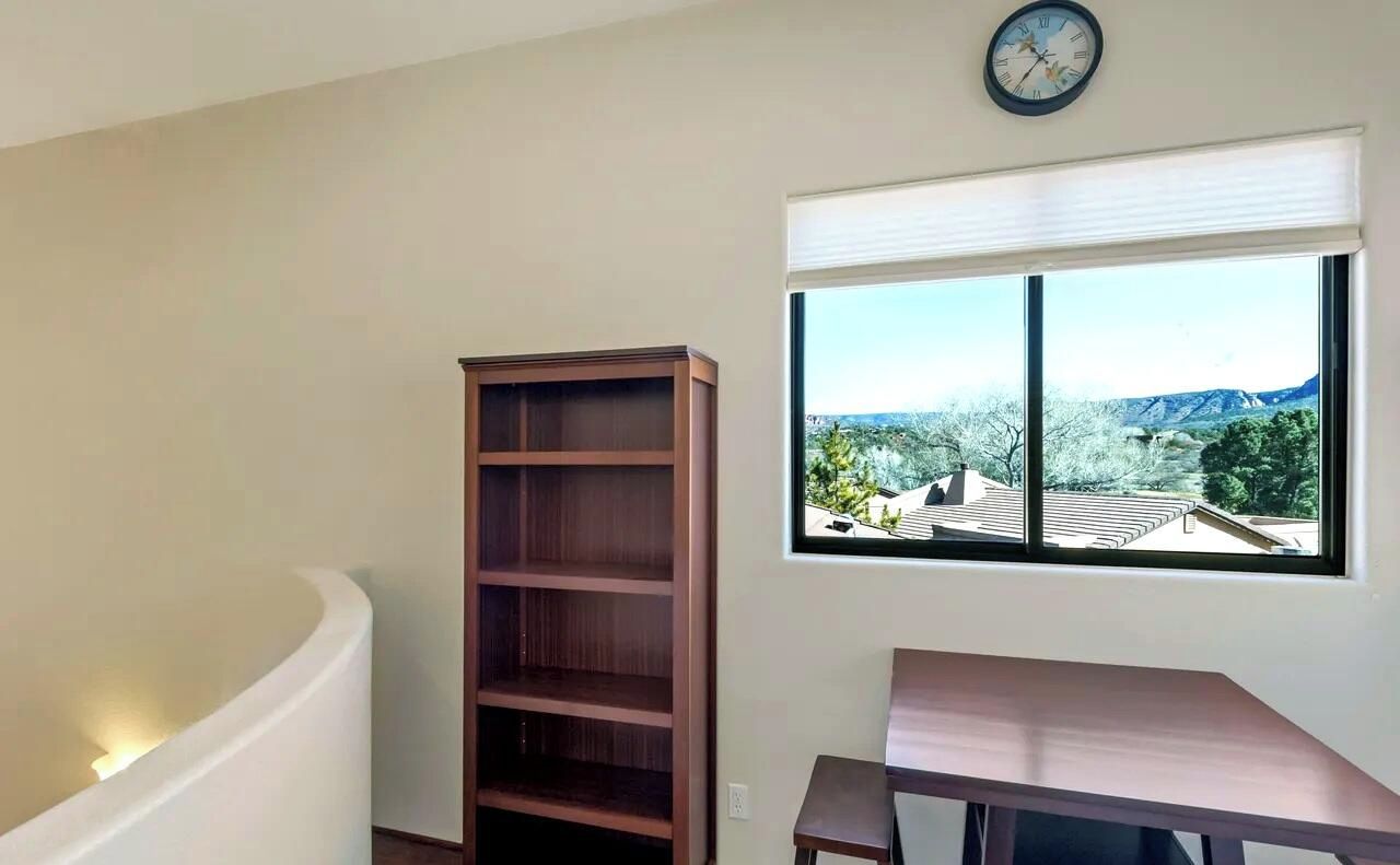 A room with a built-in bookshelf, a window with a view of a mountain and houses, and a clock on the wall.