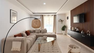 Een moderne woonkamer met een hoekbank, een glazen salontafel, een hanglamp, een flatscreen televisie bevestigd aan een houten wandpaneel en een potplant.
