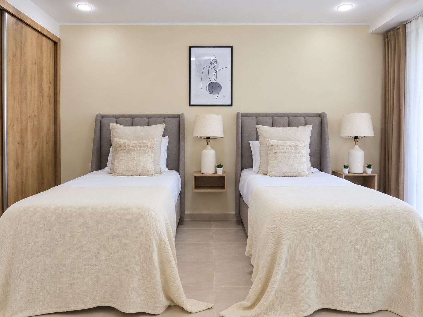 A hotel room with two beds, each with a grey headboard, white bedding, and bedside lamps. There is a framed artwork on the wall above the beds.