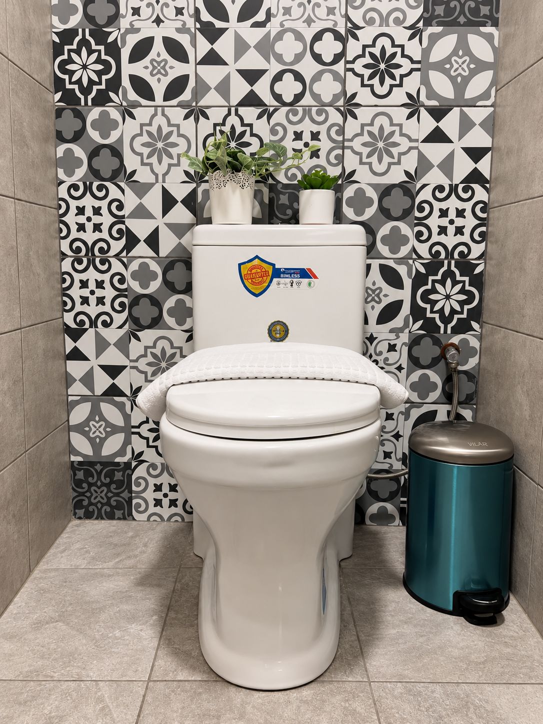 A toilet with a white lid, a toilet paper holder, and a trash can in a bathroom with patterned tiles.