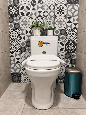 Valkoinen wc-istuimen kansi, wc-paperiteline ja roskakori kylpyhuoneessa, jonka seinät on päällystetty kuvioiduilla laatoilla.