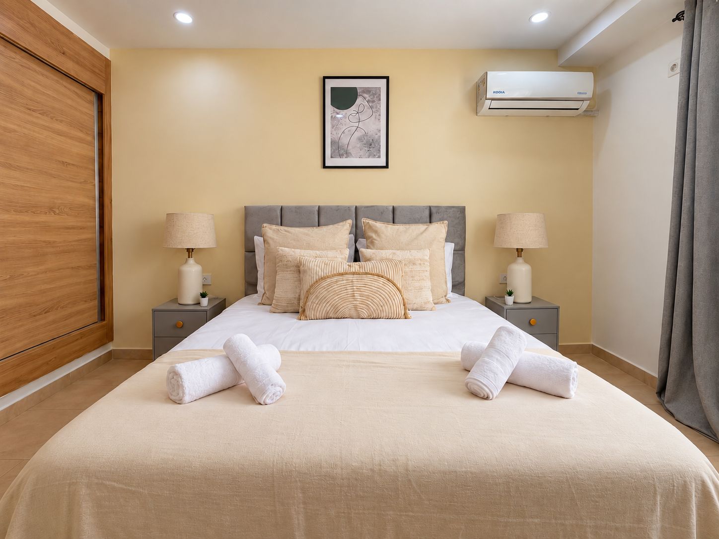 A neatly made bed with a grey headboard, two bedside tables with lamps, a framed artwork above the bed, and an air conditioning unit on the wall.