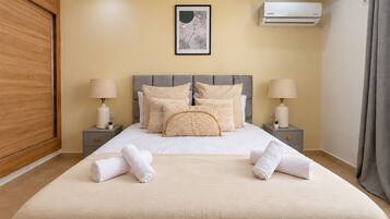 A neatly made bed with a grey headboard, two bedside tables with lamps, a framed artwork above the bed, and an air conditioning unit on the wall.