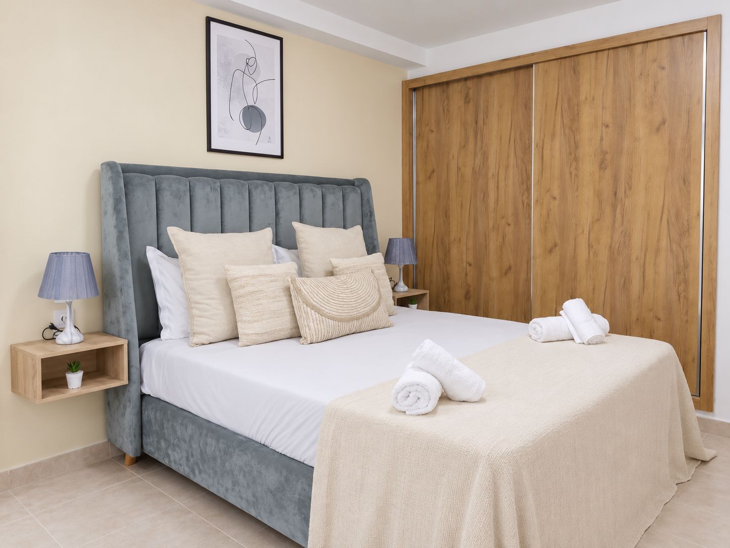 A bedroom with a bed, pillows, a bedside table with a lamp, a wooden wardrobe, and a framed picture on the wall.