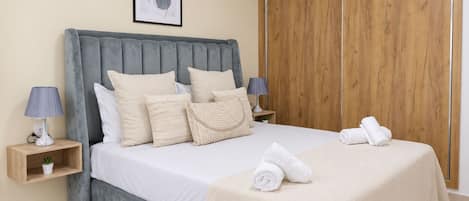 Un dormitorio con cama, almohadas, una mesita de noche con lámpara, un armario de madera y un cuadro en la pared.