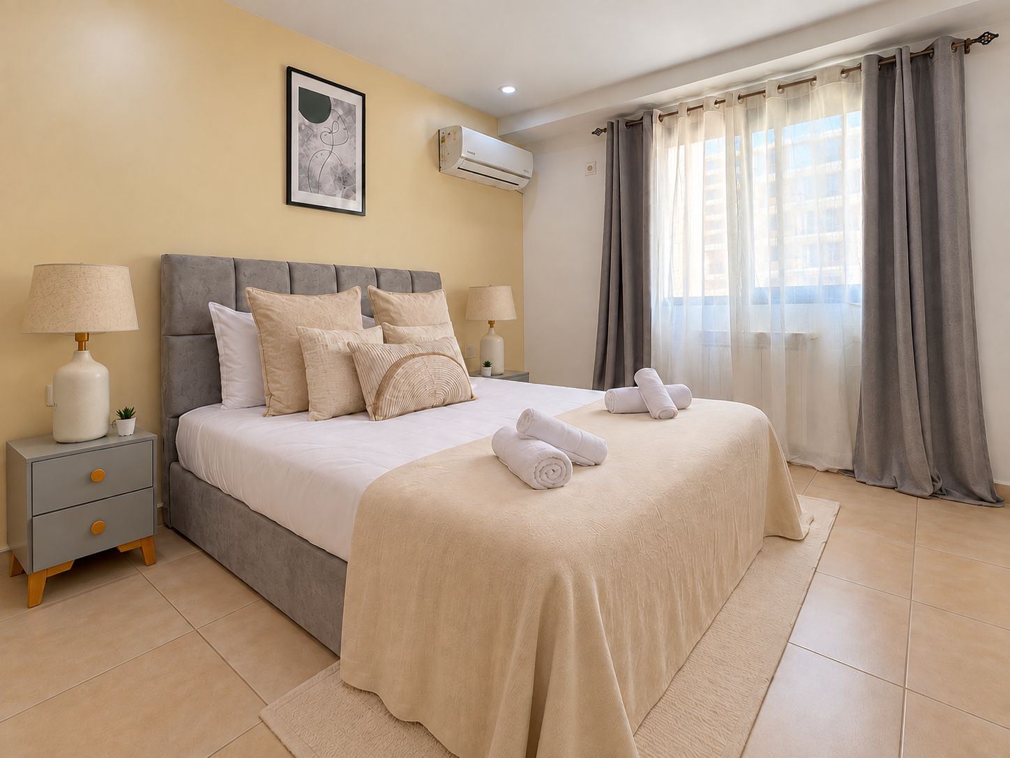 A hotel room with a bed, two bedside tables, a wall-mounted air conditioner, and a window with curtains.