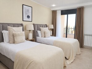 A hotel room with two beds, a window with curtains, a radiator, and a framed artwork on the wall.