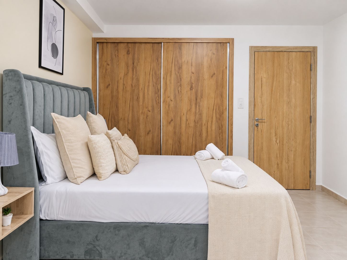 Habitación de hotel con una cama, una mesita de noche con lámpara, un cuadro enmarcado en la pared y armarios de madera.
