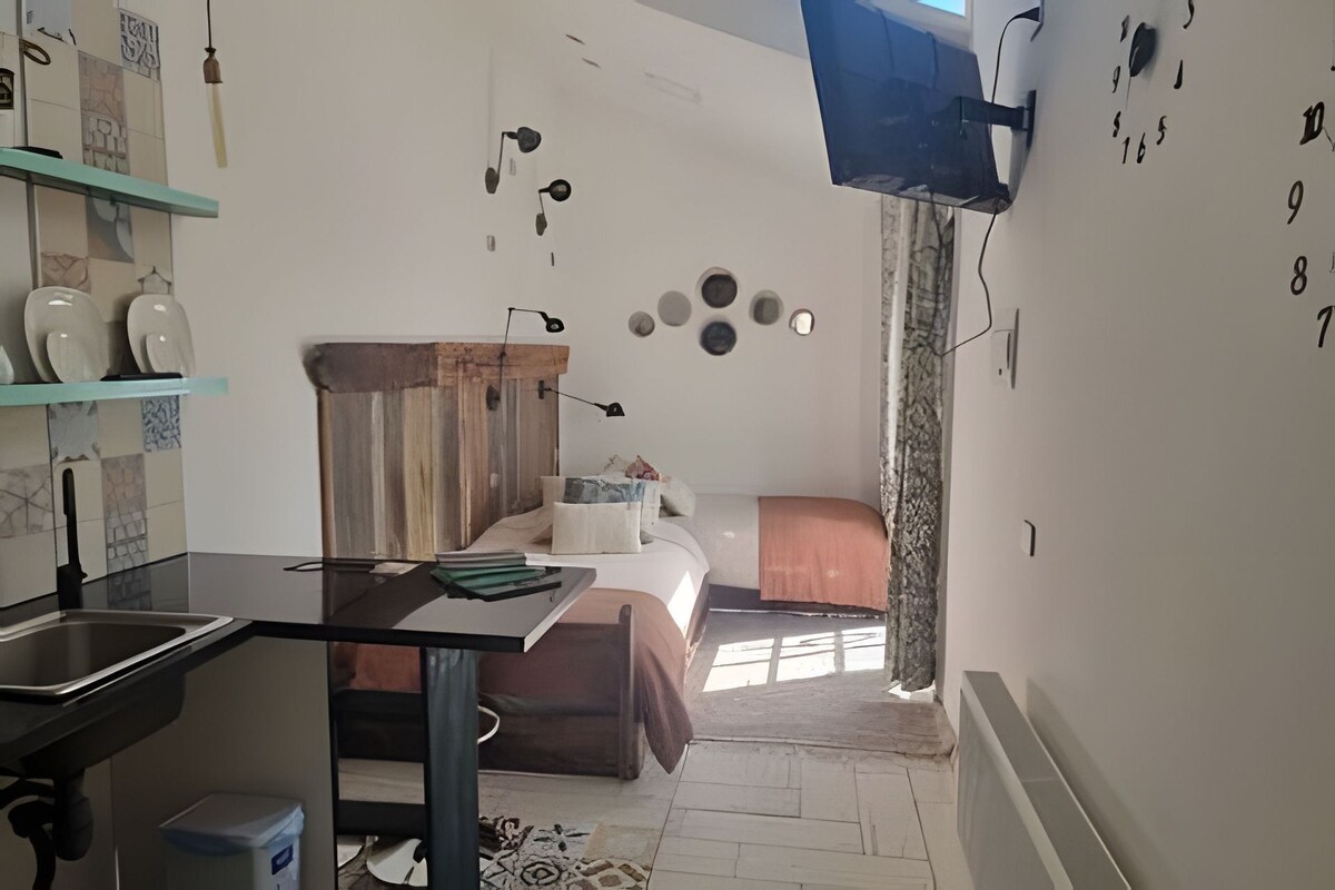 Quarto compacto com cozinha compacta, cama com cabeceira de madeira, mesa pequena e TV fixada na parede.