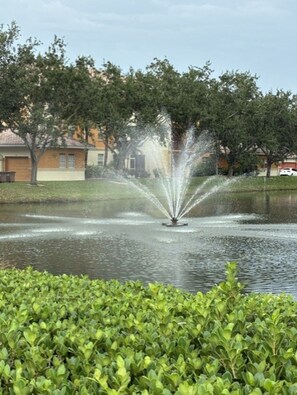A fountain in a park with a body of water, surrounded by greenery and buildings in the background. - Seasonal Rental in Cape Coral, FL (Cape Coral)