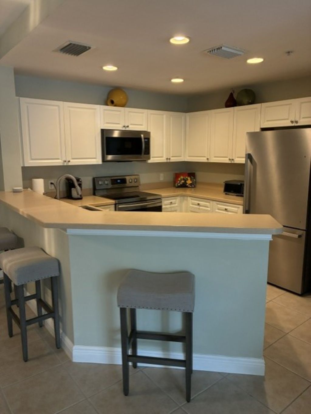 A kitchen with white cabinets, a stainless steel refrigerator, a microwave, and a stove. There are two bar stools and a small island with a sink.