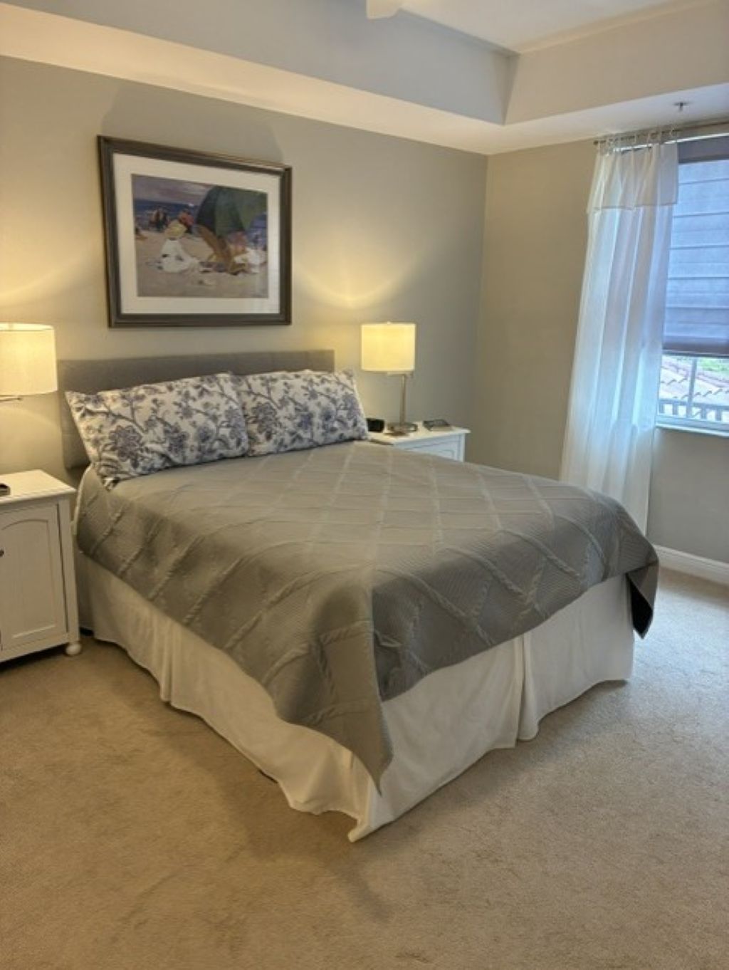 A bedroom with a bed, two bedside tables, a nightstand, a framed picture on the wall, and a window with curtains.