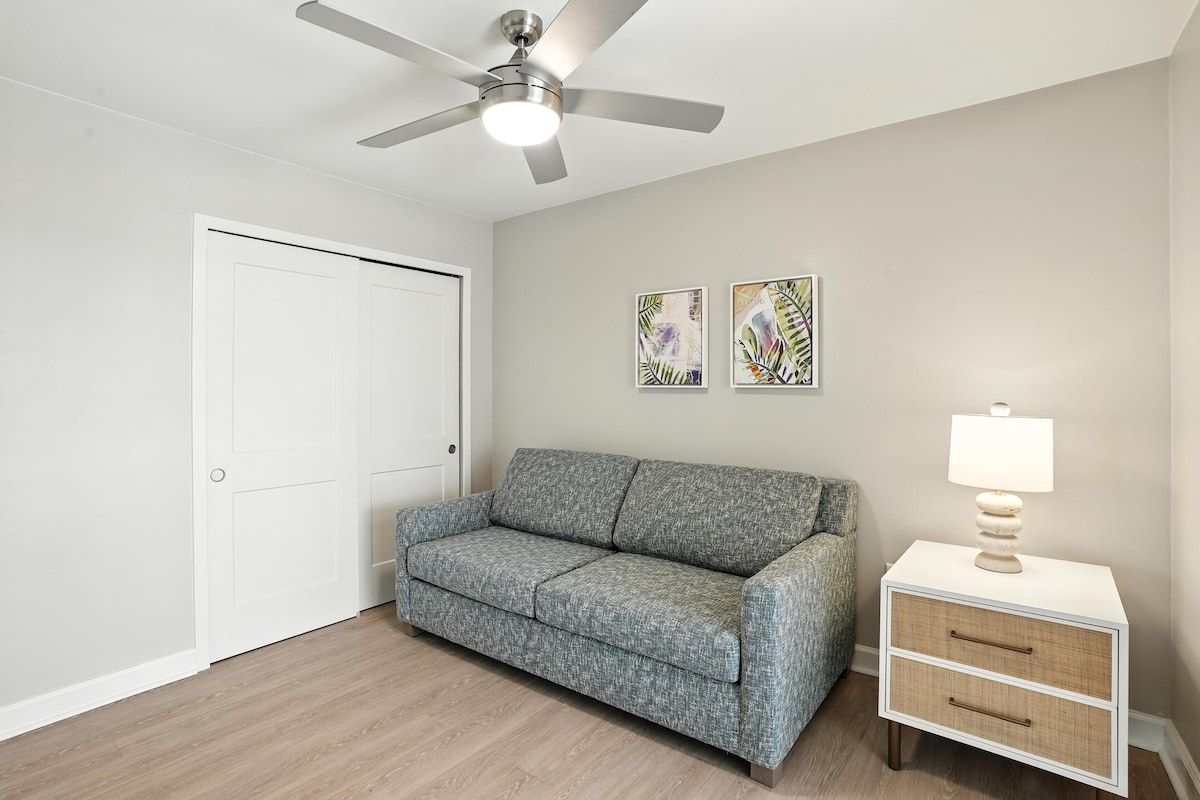 Ein Zimmer mit einem grauen Sofa, einem wei ßen Nachttisch mit Lampe und zwei abstrakten Bildern an der Wand.