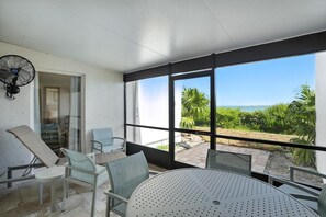 Un balcón soleado con mesa y sillas, un ventilador y vistas a zonas verdes y agua.
