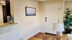 A hallway with a white door, a framed picture on the wall, a tall vase on a shelf, and a potted plant.