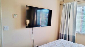 Quarto de hotel com televisão de tela plana na parede, cama com roupa de cama branca, janela com cortinas e tomada na parede.