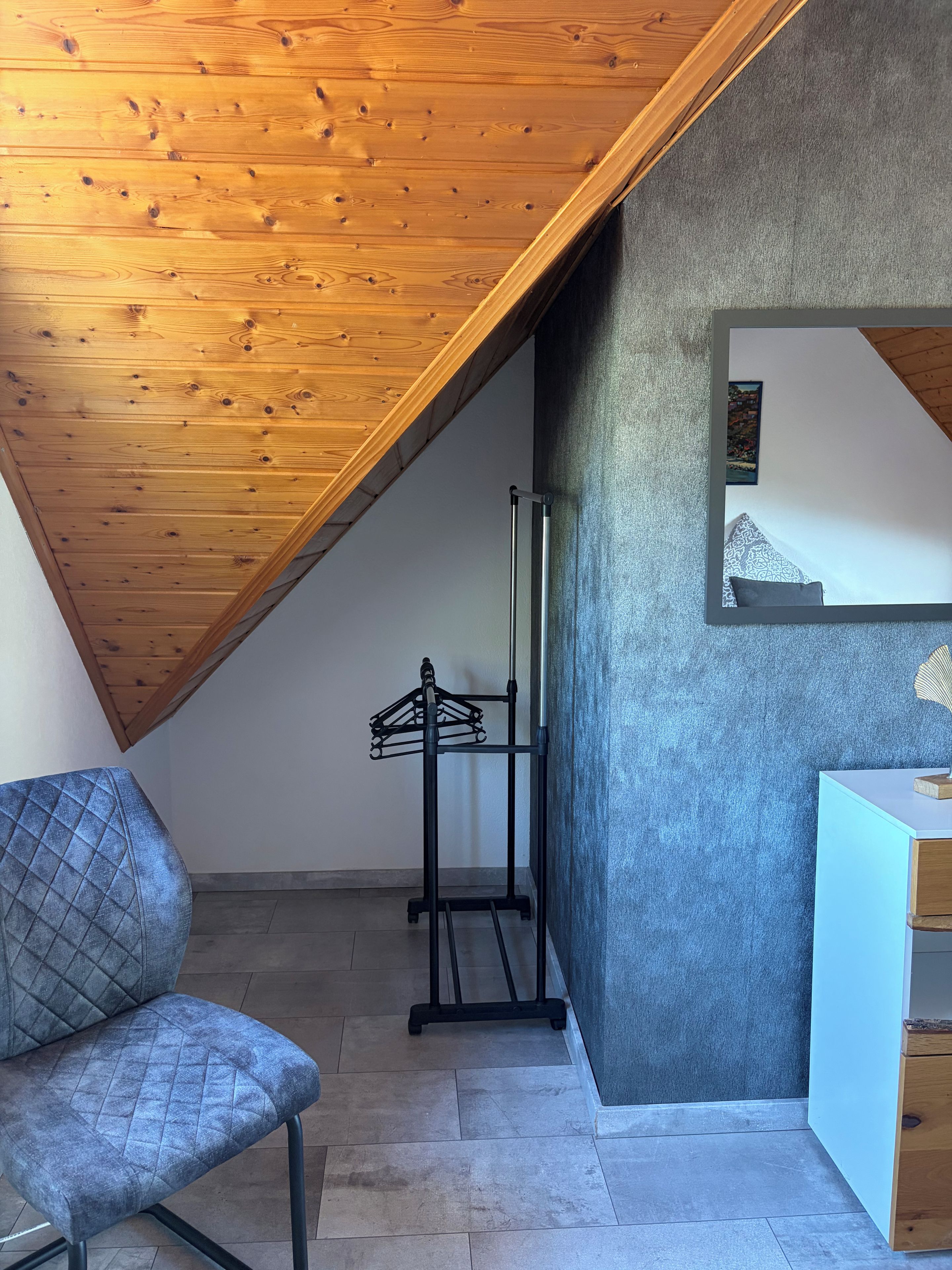 Une pièce avec un plafond en pente, un lit avec une tête de lit, une chaise, une petite table et un miroir fixé au mur.