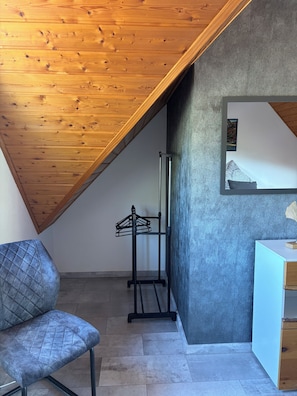 Una habitación con techo inclinado, una cama con cabecera, una silla, una mesita y un espejo instalado en la pared.