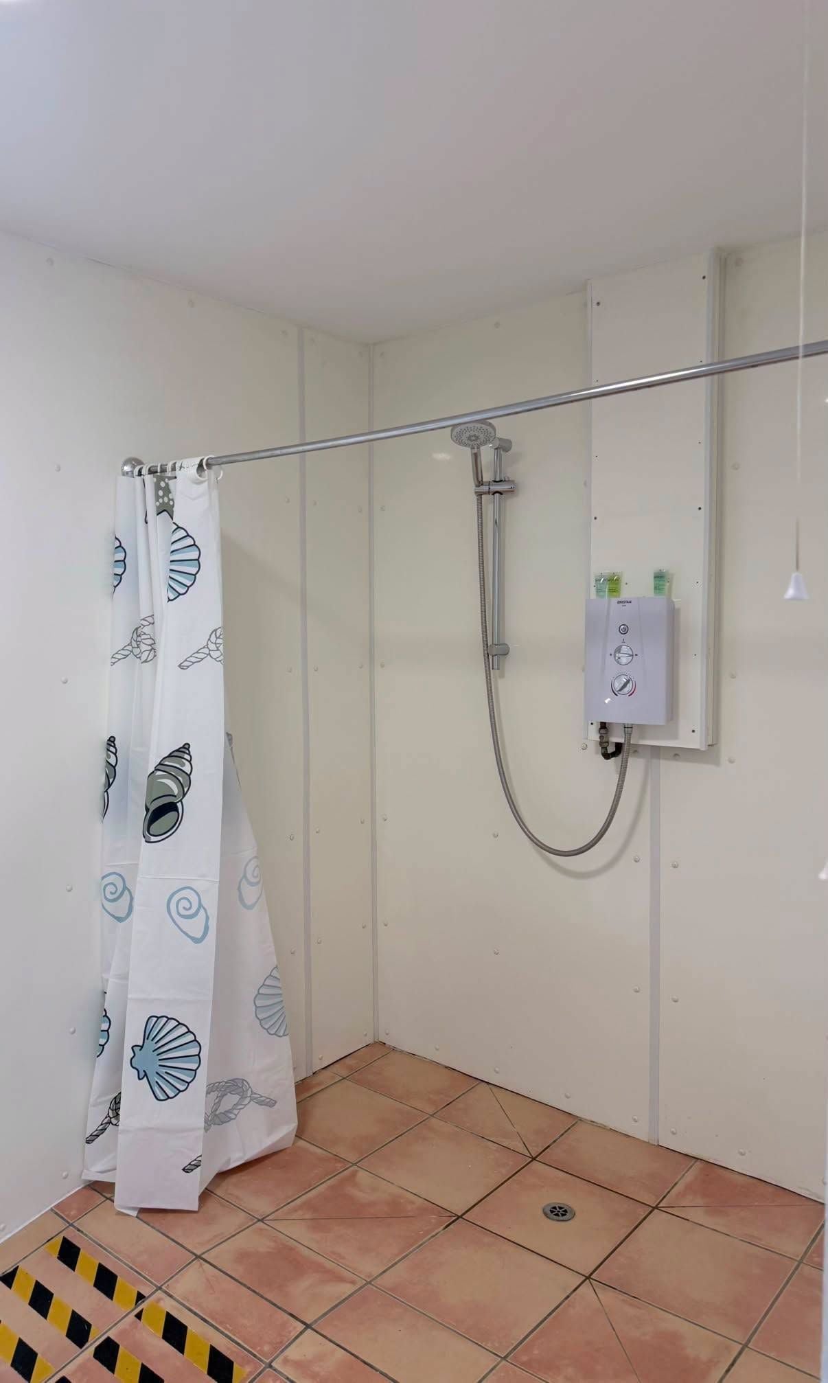 A small bathroom with a shower, a white curtain with shell patterns, and a tiled floor.