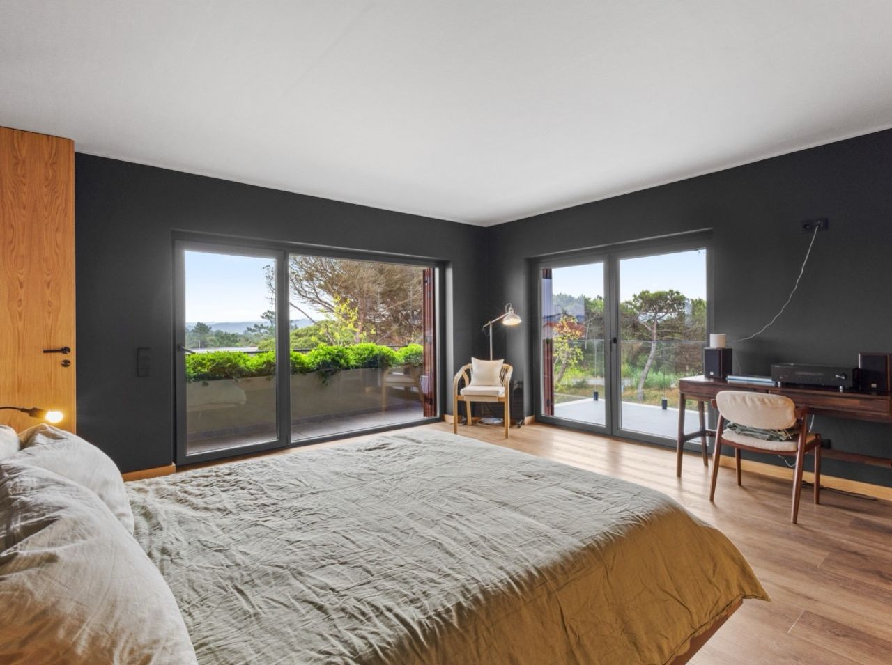 A modern bedroom with a large bed, a desk, and a chair, featuring a view through glass doors.