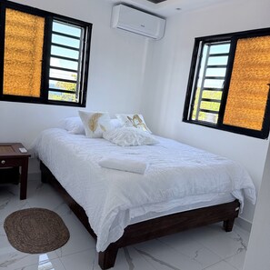 A bedroom with a bed, pillows, a bedside table, an air conditioning unit, and a window with a view.