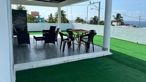 Eine Terrasse im Freien mit Grill, Esstisch und Stühlen, mit Blick auf eine grüne Rasenfläche und das Meer.