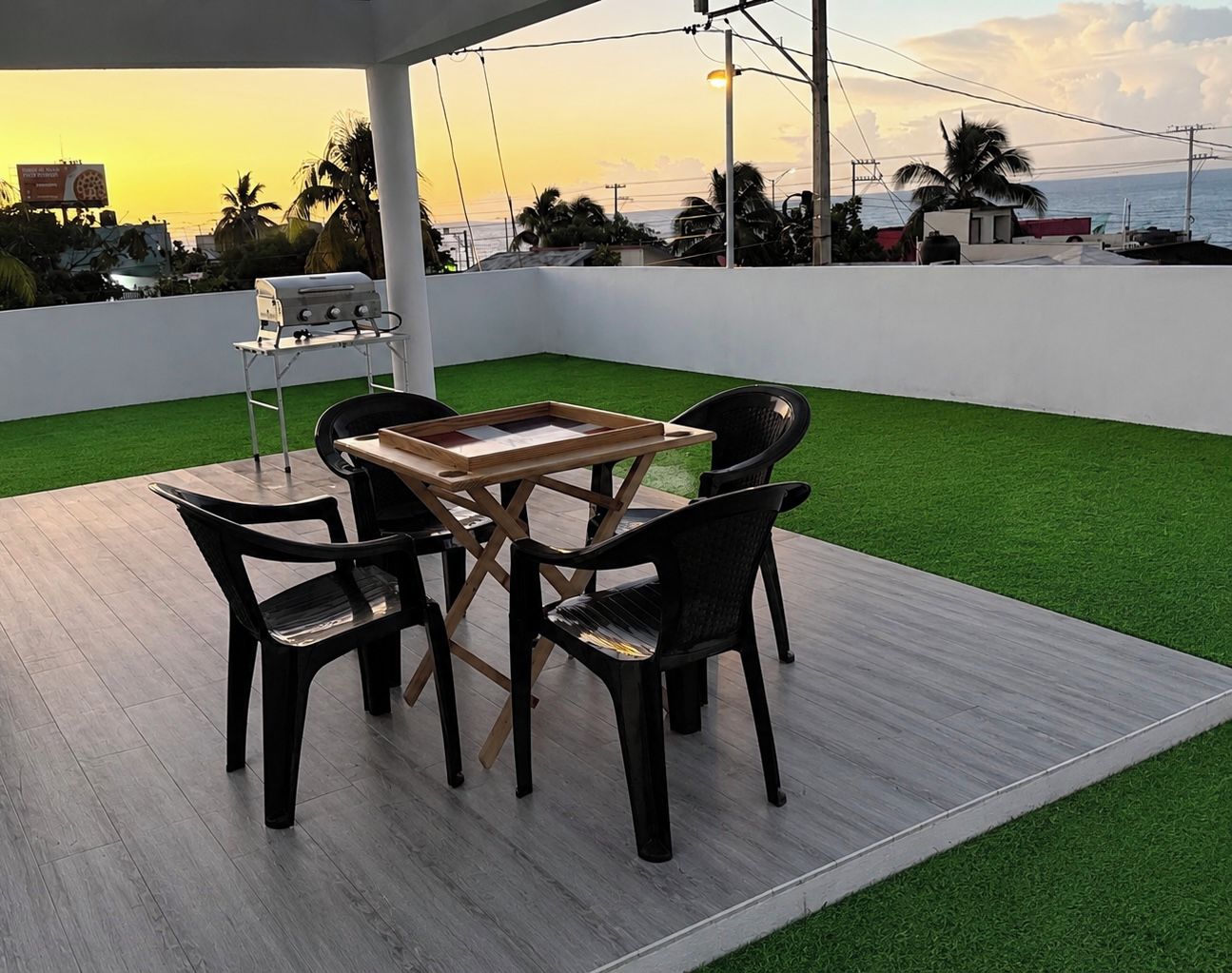 Une terrasse extérieure avec une table et des chaises, de l’herbe artificielle et une vue sur la mer au coucher du soleil.