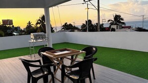 Eine Außenpatio mit Tisch, Stühlen, Kunstgras und Blick auf das Meer bei Sonnenuntergang.