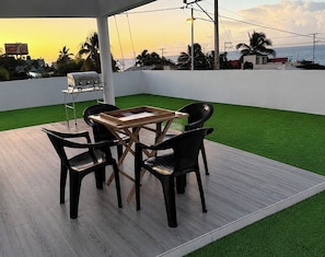 An outdoor patio with a table and chairs, artificial grass, and a view of the sea at sunset. - Sunrise Ocean Views - Private Rooftop - BBQ - Near SDQ (Santo Domingo Este)