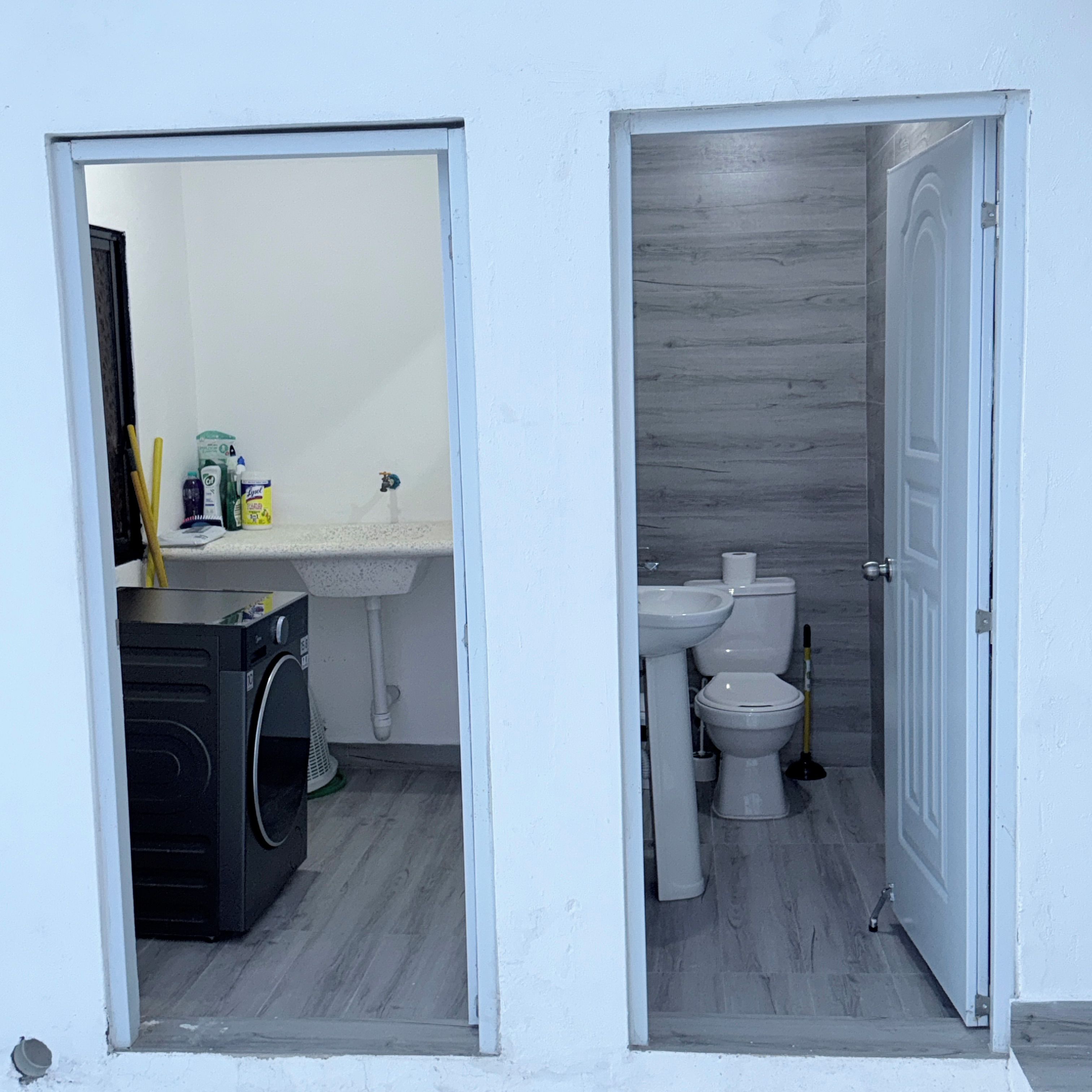 A bathroom with a toilet, sink, and mirror; a laundry area with a washing machine; and a door leading to another room.