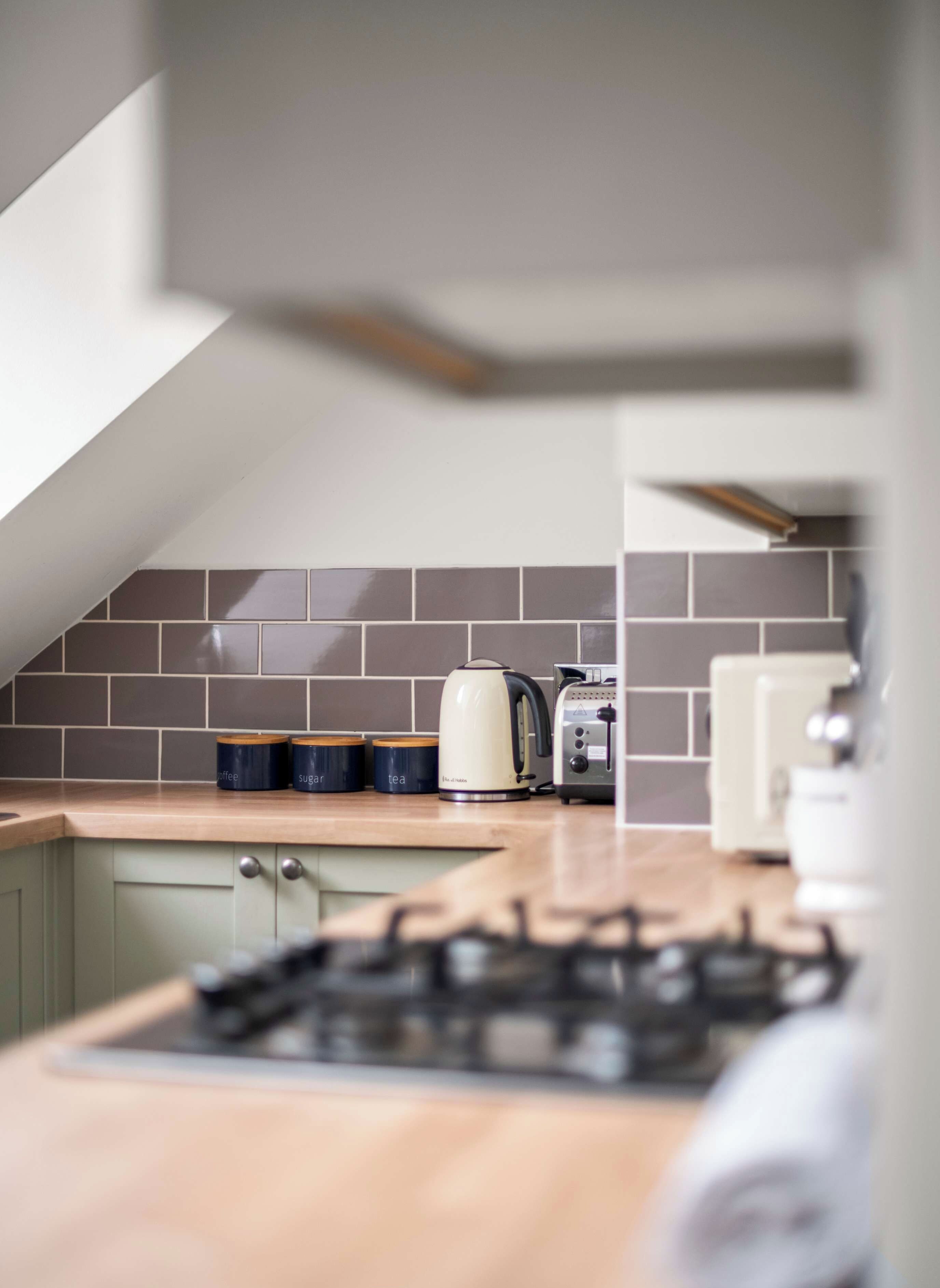 A modern kitchen counter with a built-in stovetop features a cream-colored electric kettle, a toaster, and matching storage canisters against a gray subway tile backsplash.