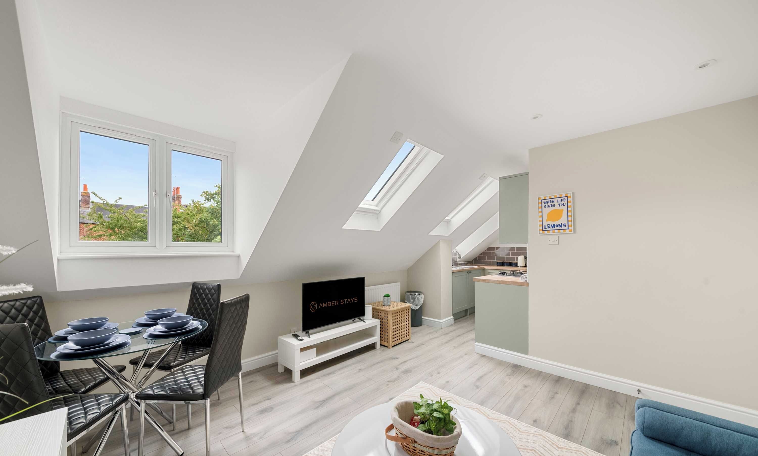 An open-concept living space features a glass dining table set for four, a television on a white stand under a sloped ceiling, and a kitchen area .
