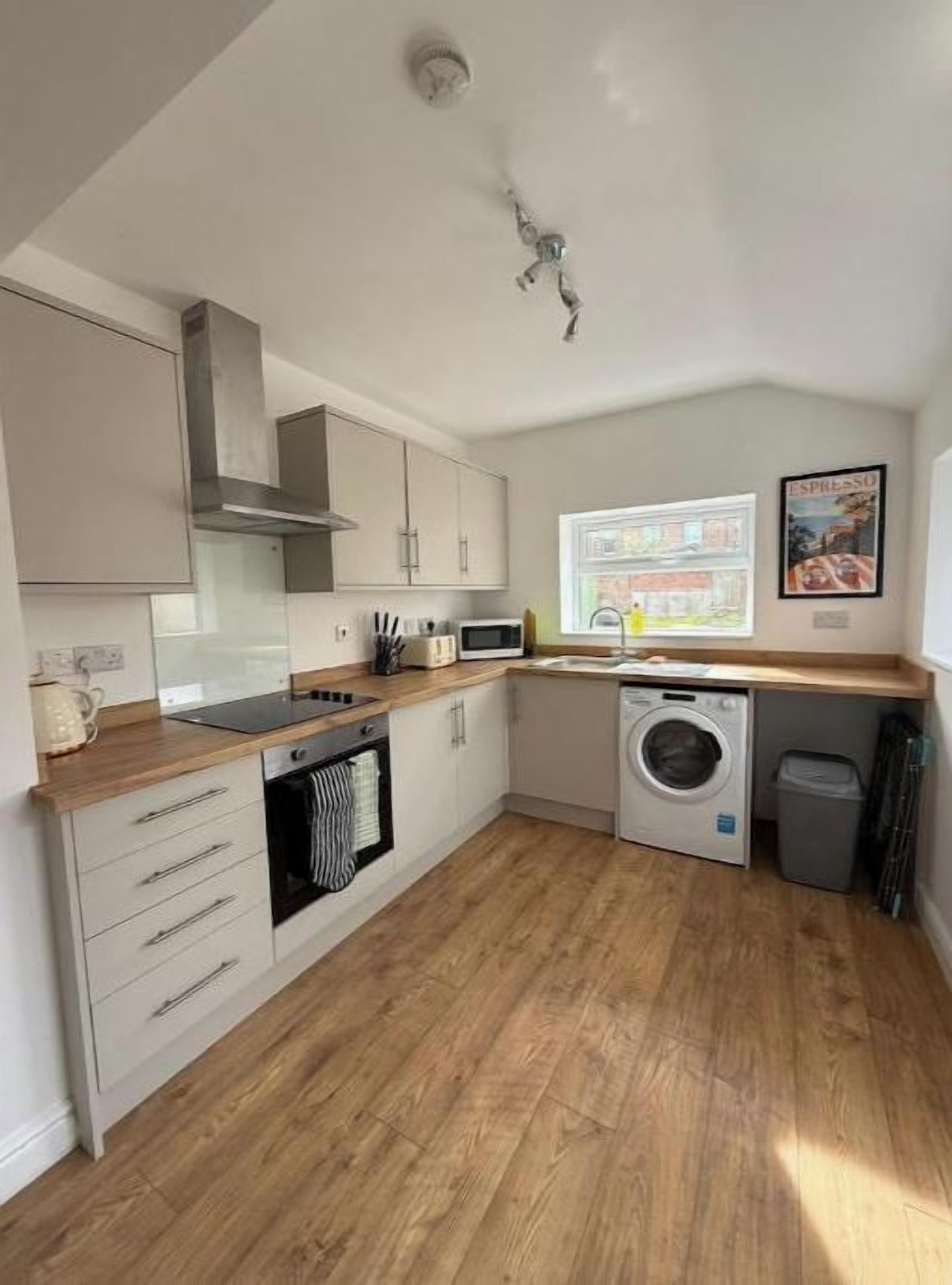 A compact kitchen with a microwave, oven, and washing machine, wooden flooring, and a window with a view.