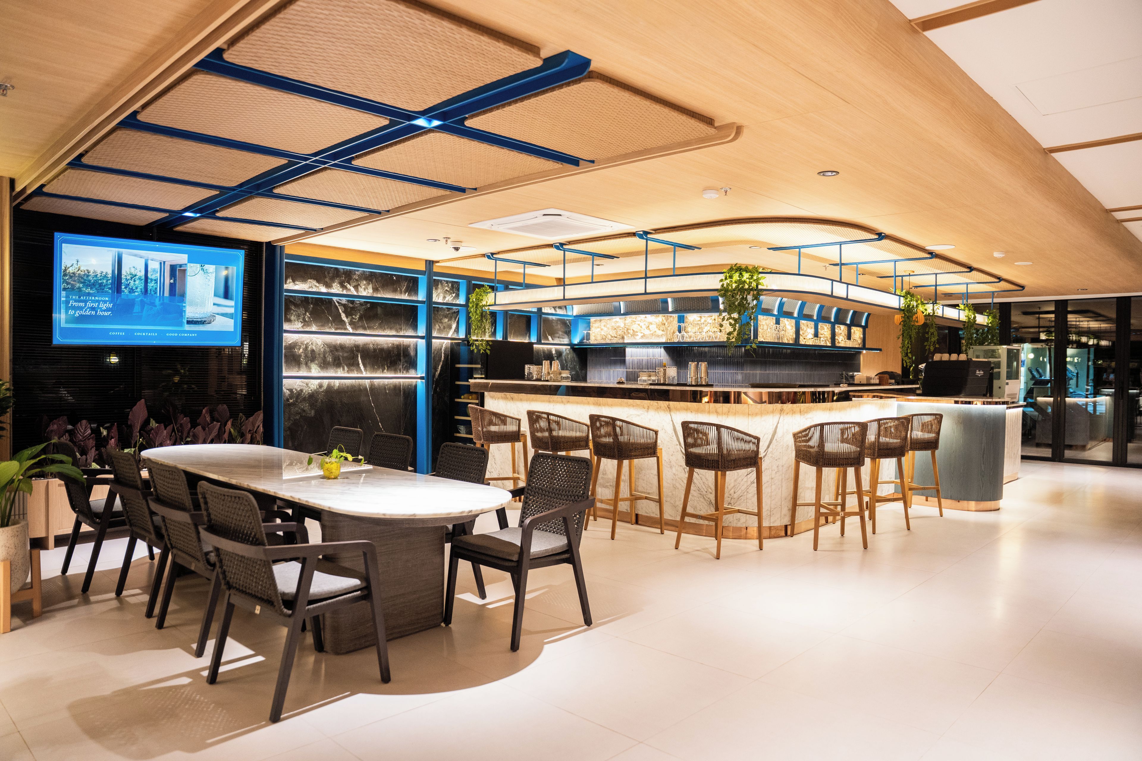 Un área de bar moderna con una barra curva, diversas opciones de asientos y una pantalla grande mostrando contenido.