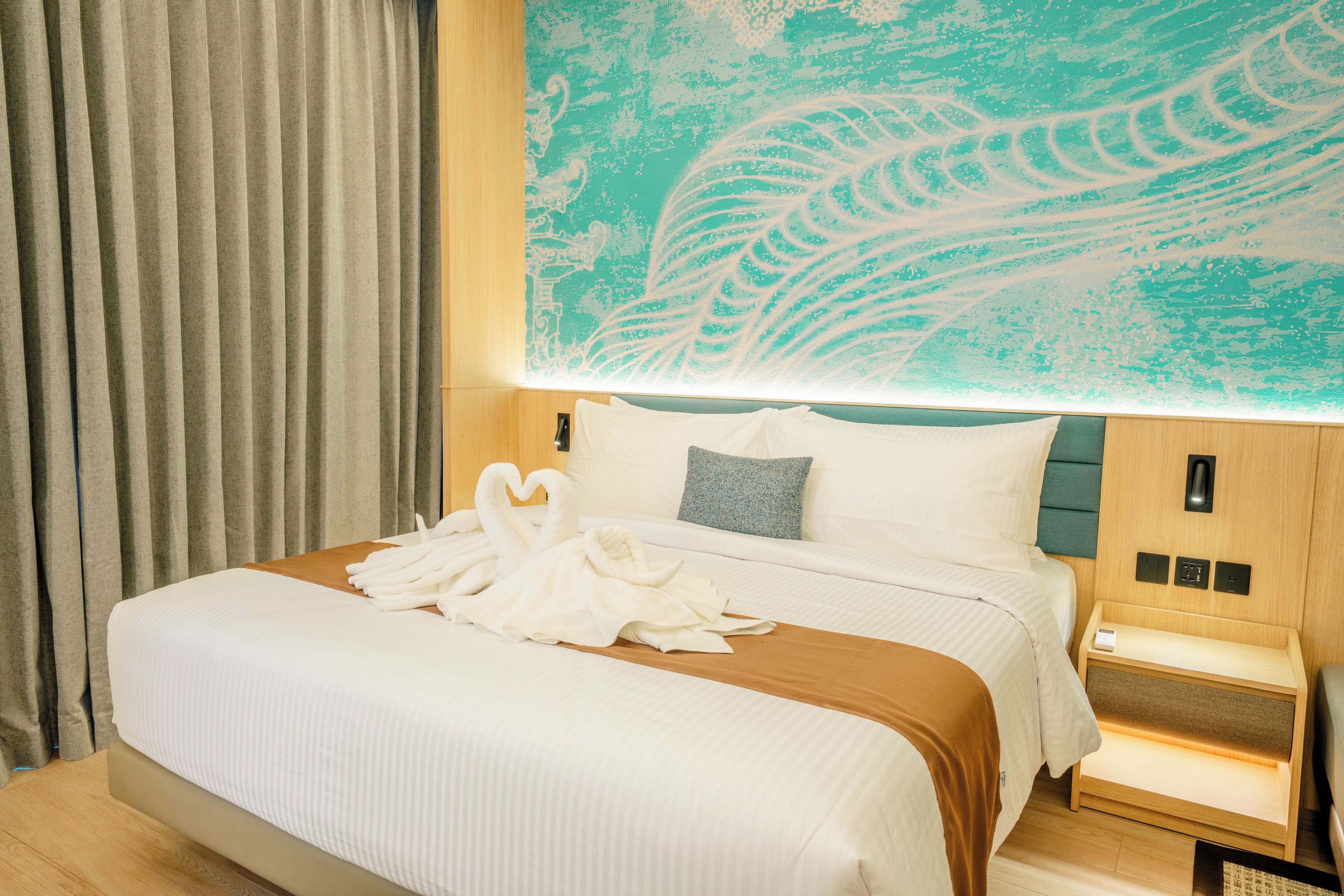 Una habitación de hotel con una cama grande, cabecera con diseño abstracto color aguamarina, una mesita de noche de madera y una lámpara fijada a la pared.
