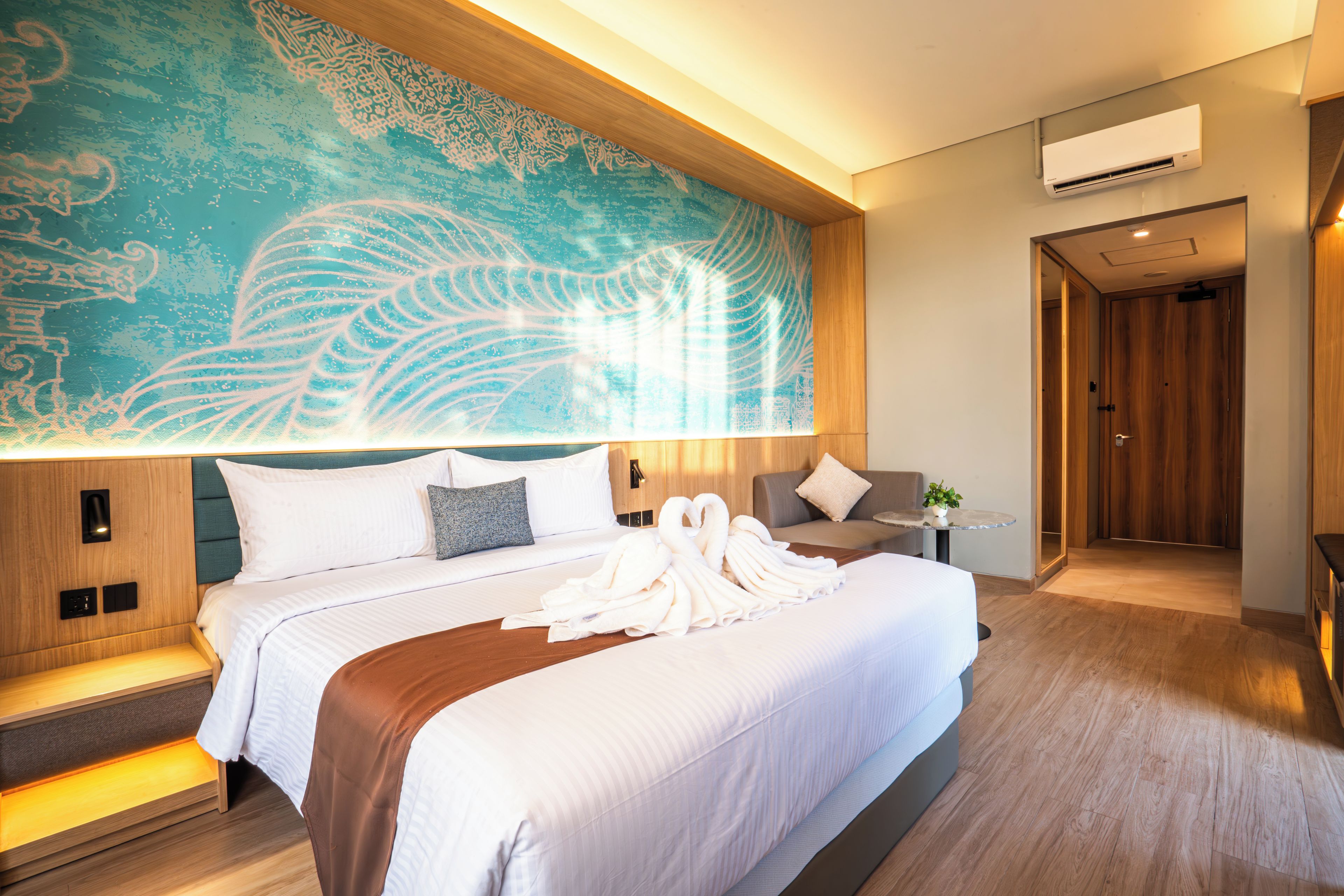 Habitación de hotel con una cama grande, un mural en la pared, una mesita de noche con una planta y una puerta que da a otra habitación.
