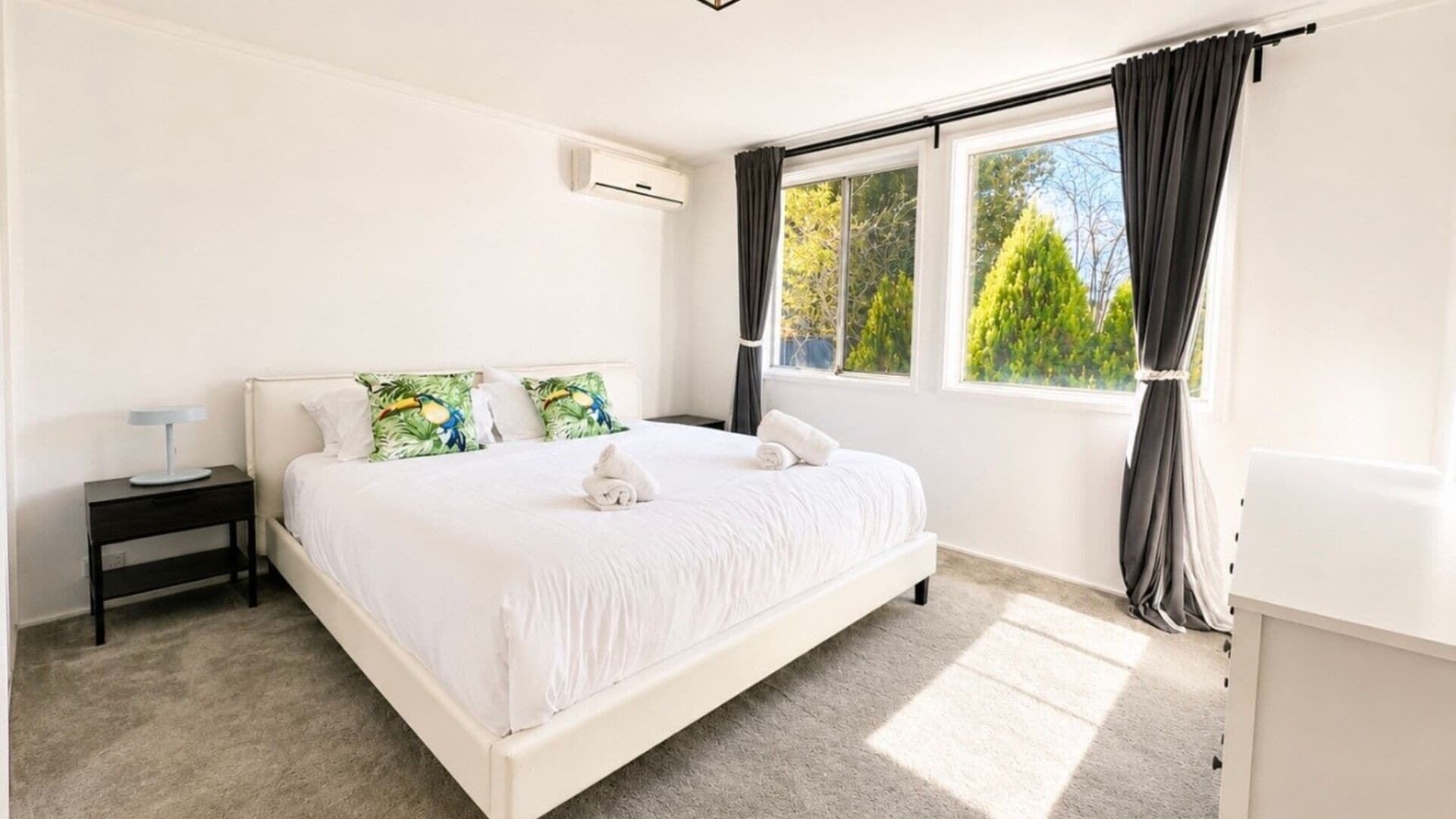 A bedroom with a bed, a nightstand, a window with curtains, and an air conditioning unit.