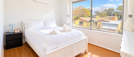 A bedroom with a bed, a nightstand, a window with a view of houses, and an air conditioning unit.