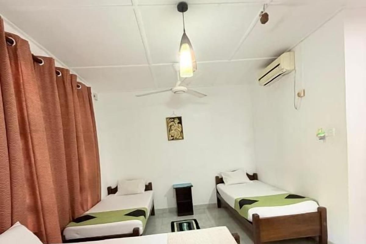 Quarto de hotel com duas camas de solteiro, ventoinha no teto, ar condicionado e uma mesinha.