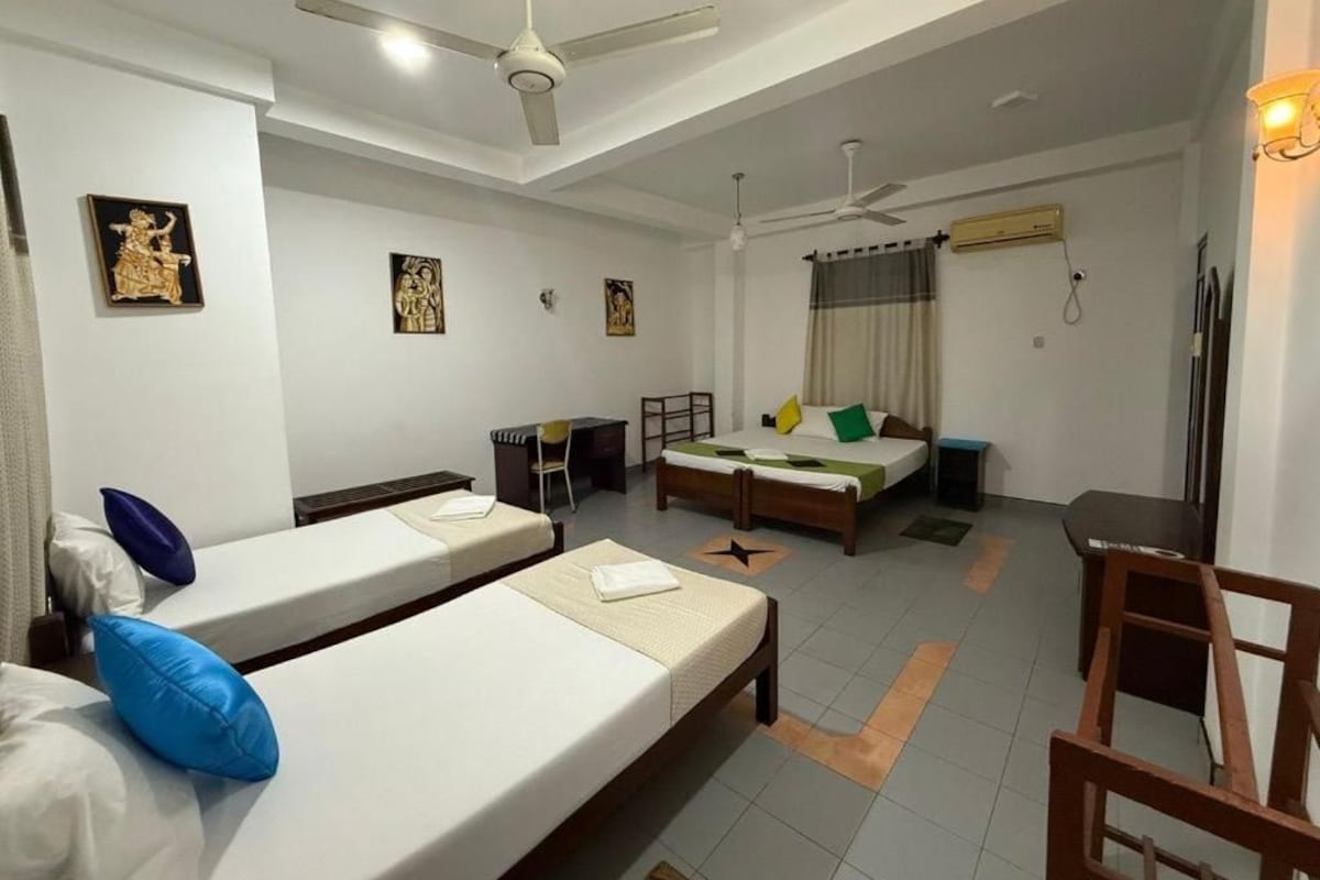 Quarto de hotel com duas camas de solteiro, uma escrivaninha, uma cadeira e um ventilador de teto.