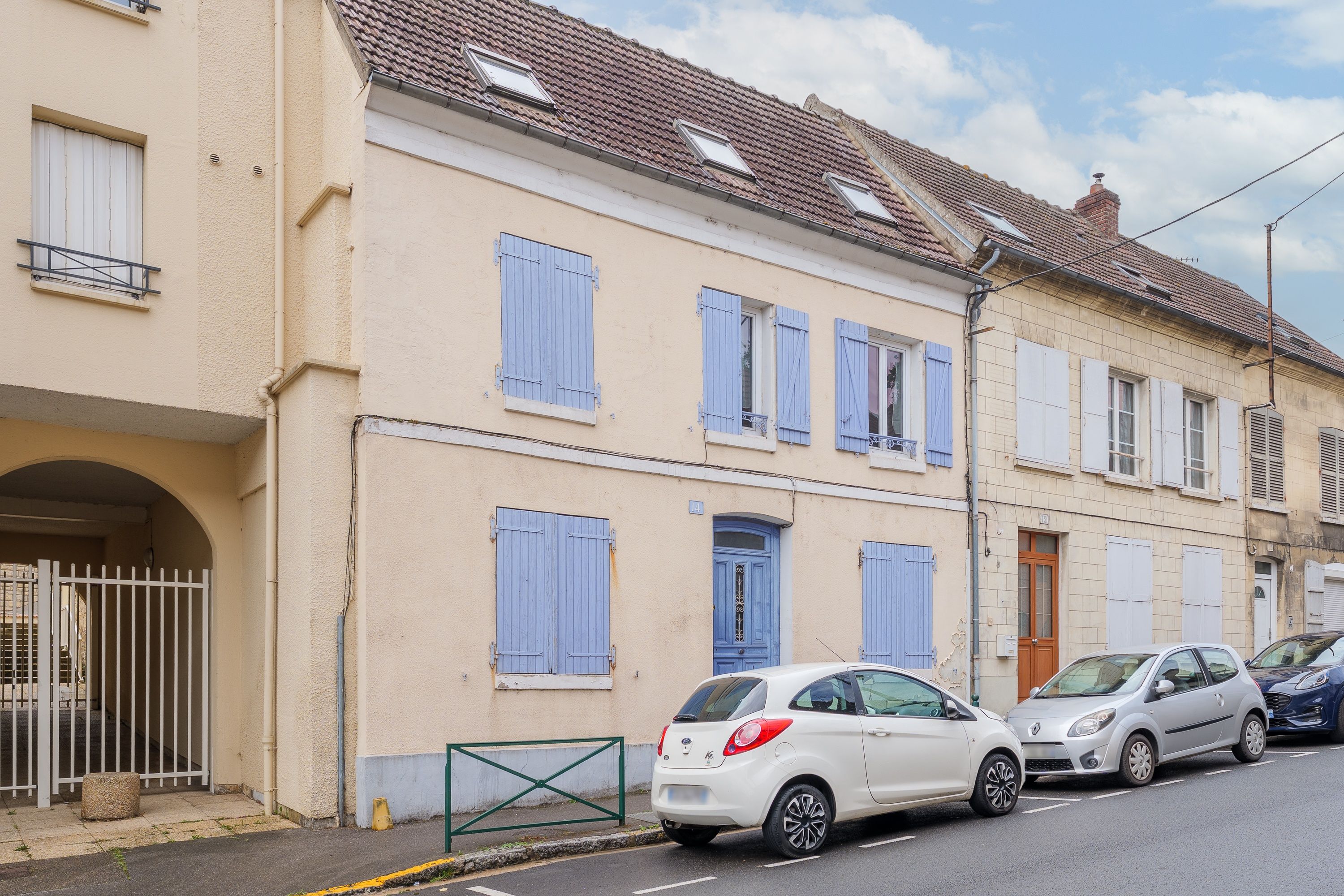 Une rue résidentielle bordée de maisons à deux étages, chacune dotée d’un balcon et d’une petite fenêtre. Des voitures sont garées le long de la rue.