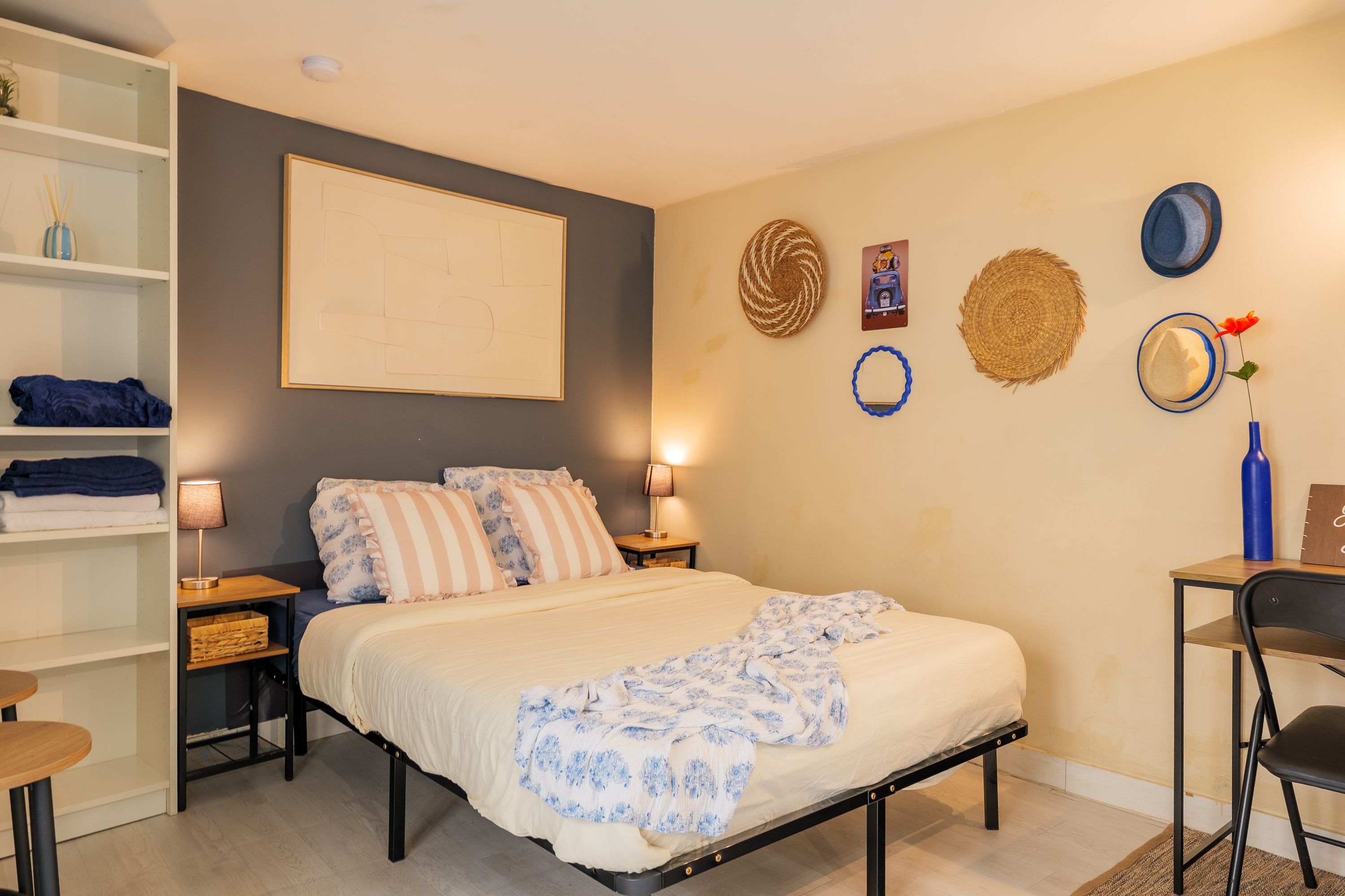 A bedroom with a bed, bedside tables, a lamp, a chair, a shelf with towels, and decorative wall items.