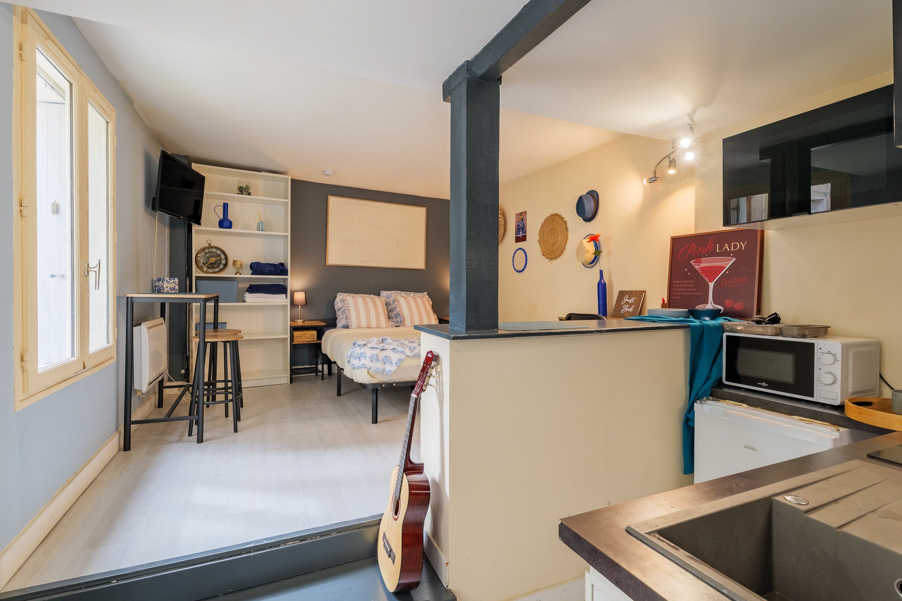 Een knus woongedeelte met een kitchenette, een bed met gestreepte lakens, een gitaar en een tv aan de muur.