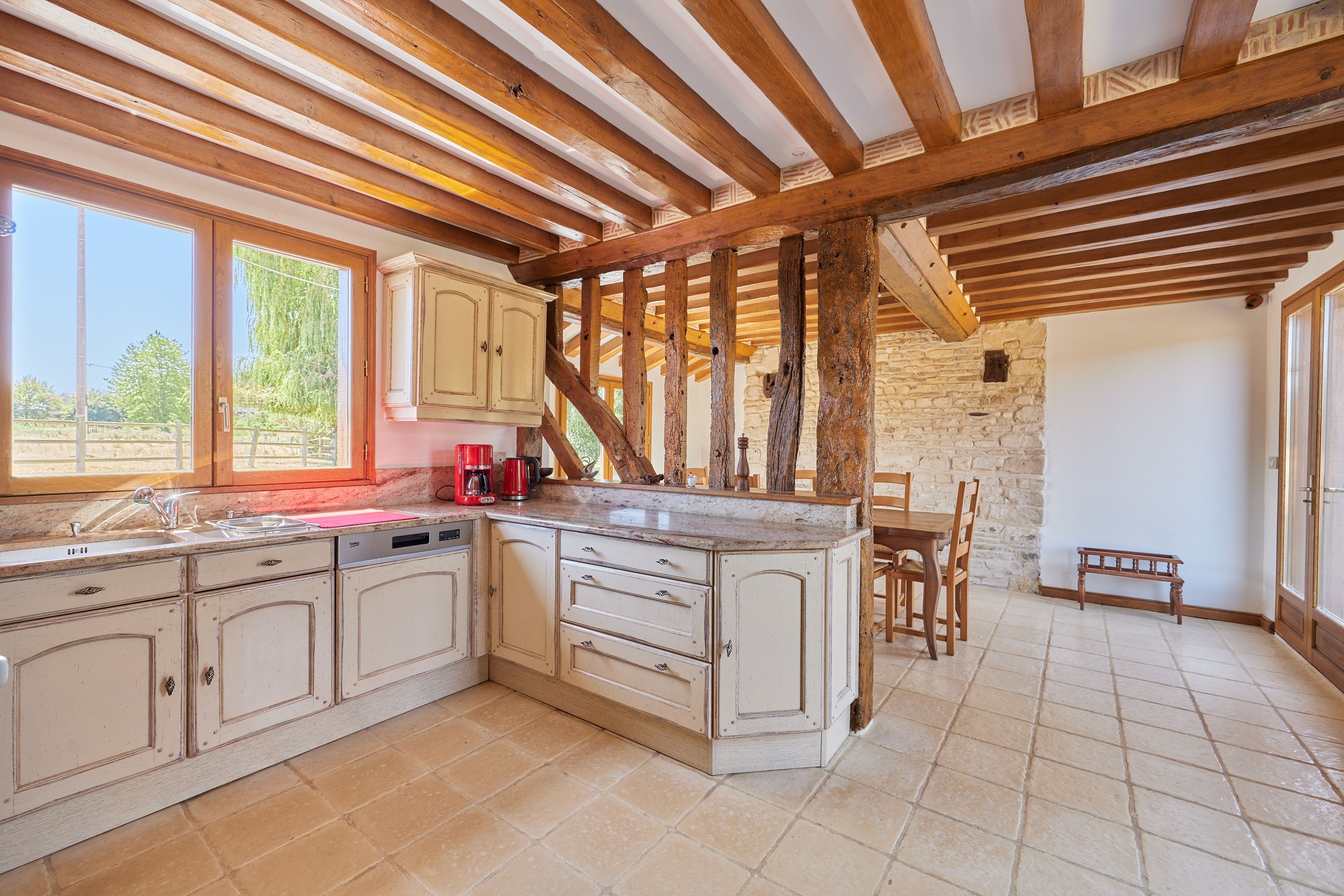 A kitchen with wooden beams, a stone wall, and a view of greenery outside.