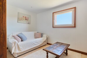 A room with a white sofa, a small table with a patterned top, a window with a wooden frame, and a wall-mounted picture. - Maison du Chemin des Vignes - charme normand (L'Oudon)