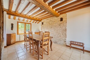 A dining area with a wooden table and chairs, a stone wall, and a window with a view of greenery. - Maison du Chemin des Vignes - charme normand (L'Oudon)