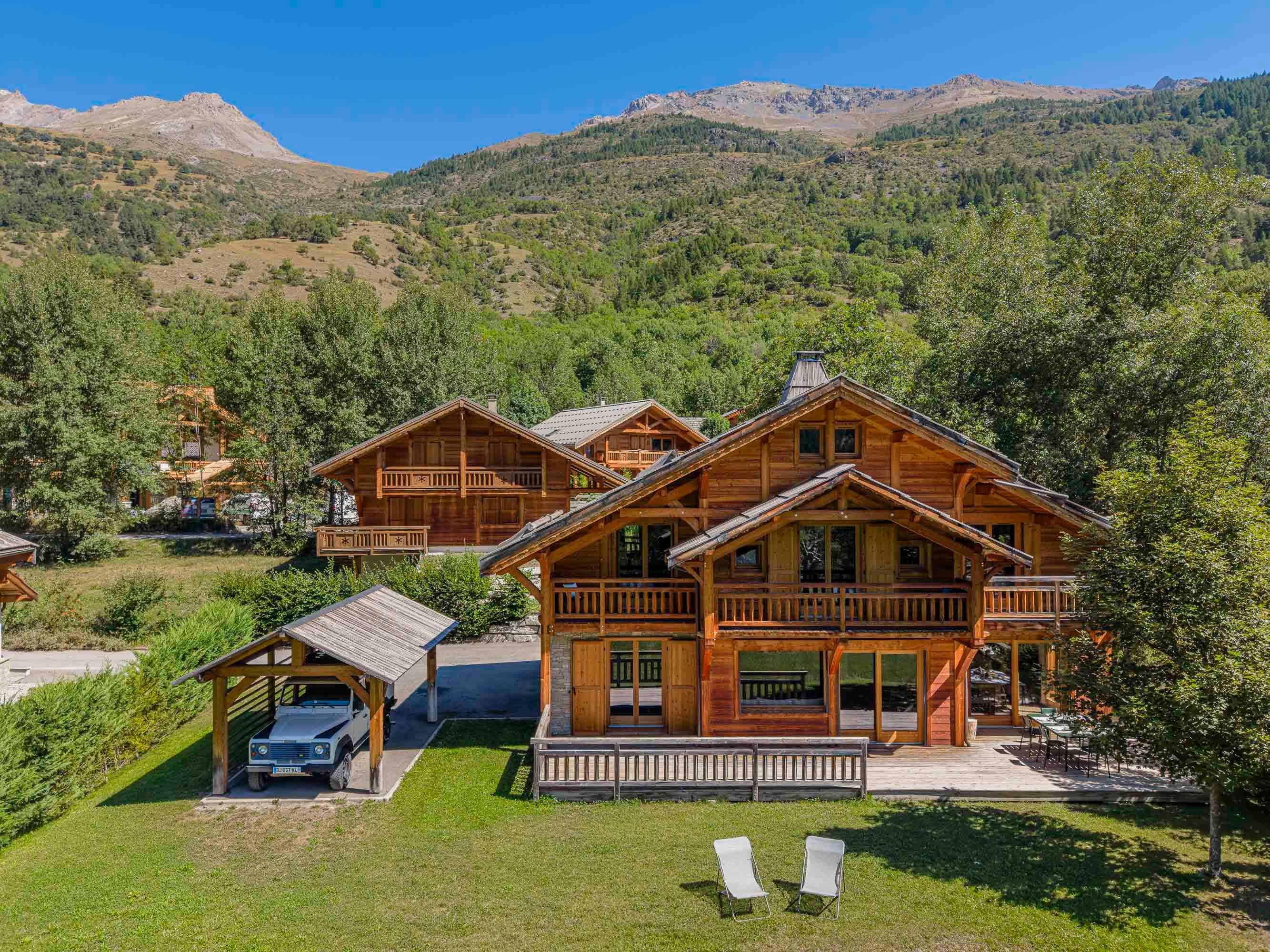 A wooden cabin with a covered carport, a patio with outdoor seating, and a view of a mountainous landscape.